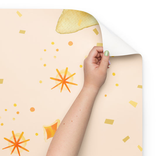 Hand peeling the corner of a peach-colored wrapping paper sheet with watercolor-style yellow and orange starburst and confetti patterns.