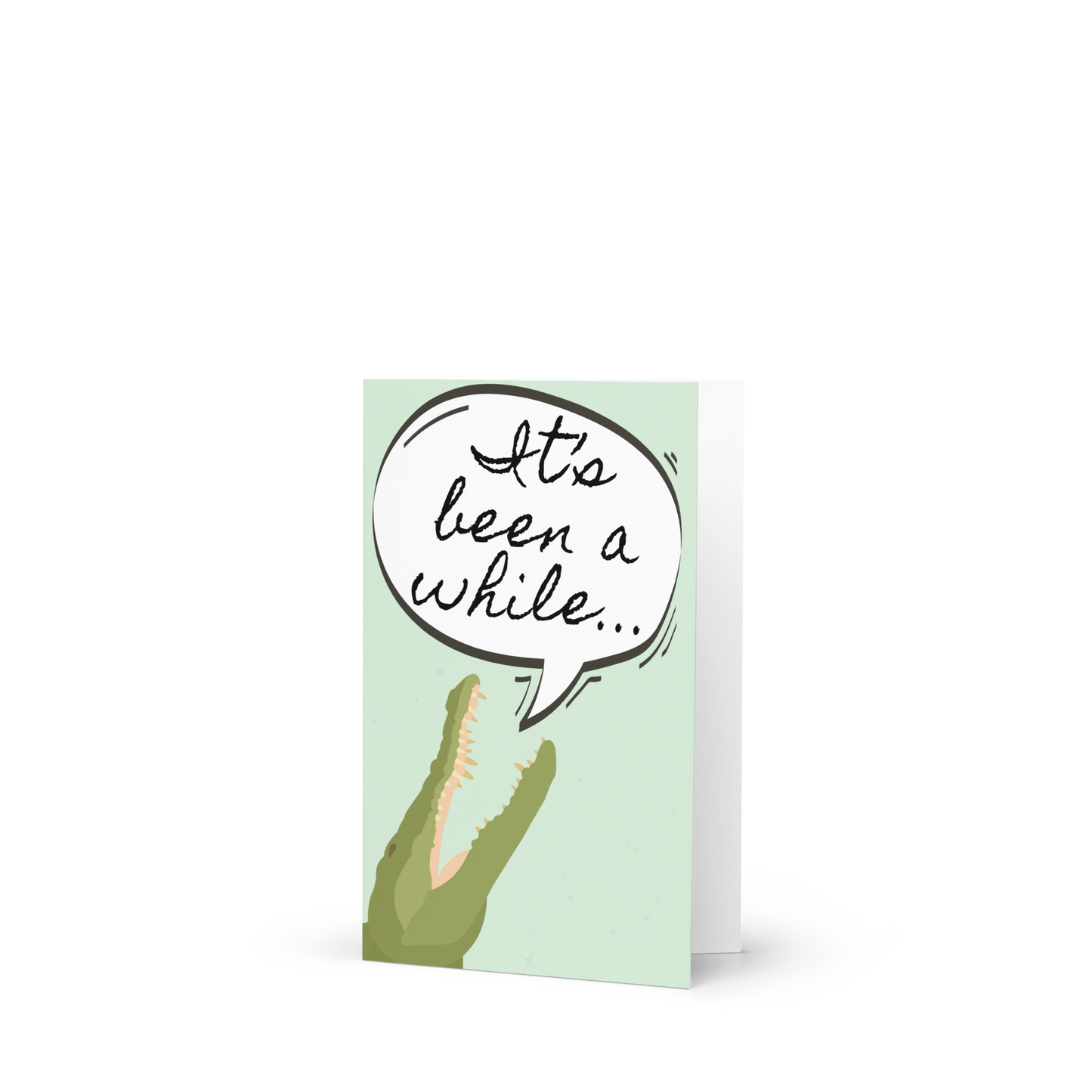 Greeting card with a crocodile and speech bubble saying 'It's been a while' on a light green background