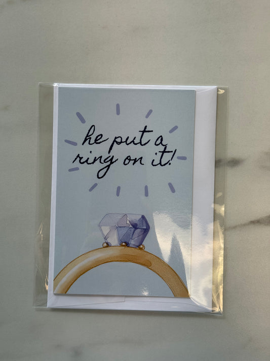Greeting card with a diamond ring illustration and text 'he put a ring on it!' on a light gray background.