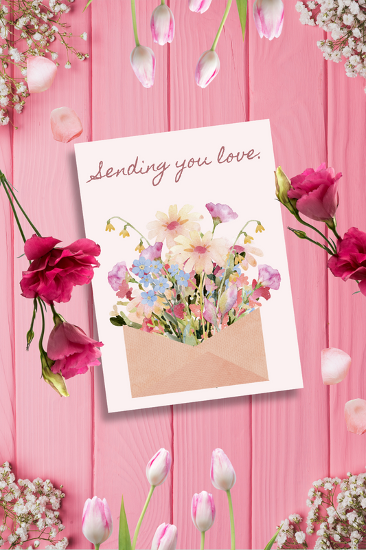 Greeting card with floral design and 'Sending you love' text on a pink wooden background with flowers.