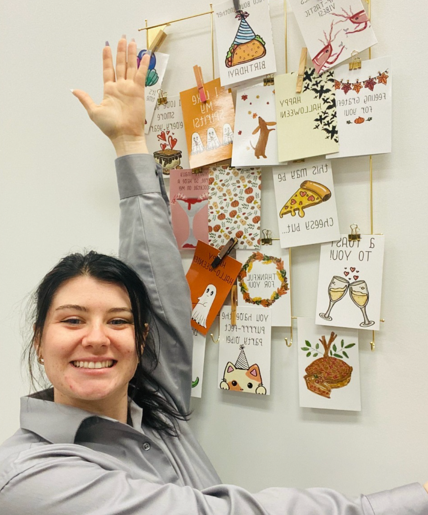 Smiling woman raising her hand in front of a display of illustrated greeting cards clipped to a wall, featuring playful designs and handwritten-style messages.