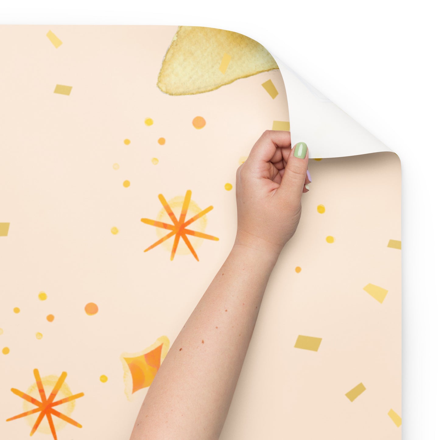Hand peeling the corner of a peach-colored wrapping paper sheet with watercolor-style yellow and orange starburst and confetti patterns.