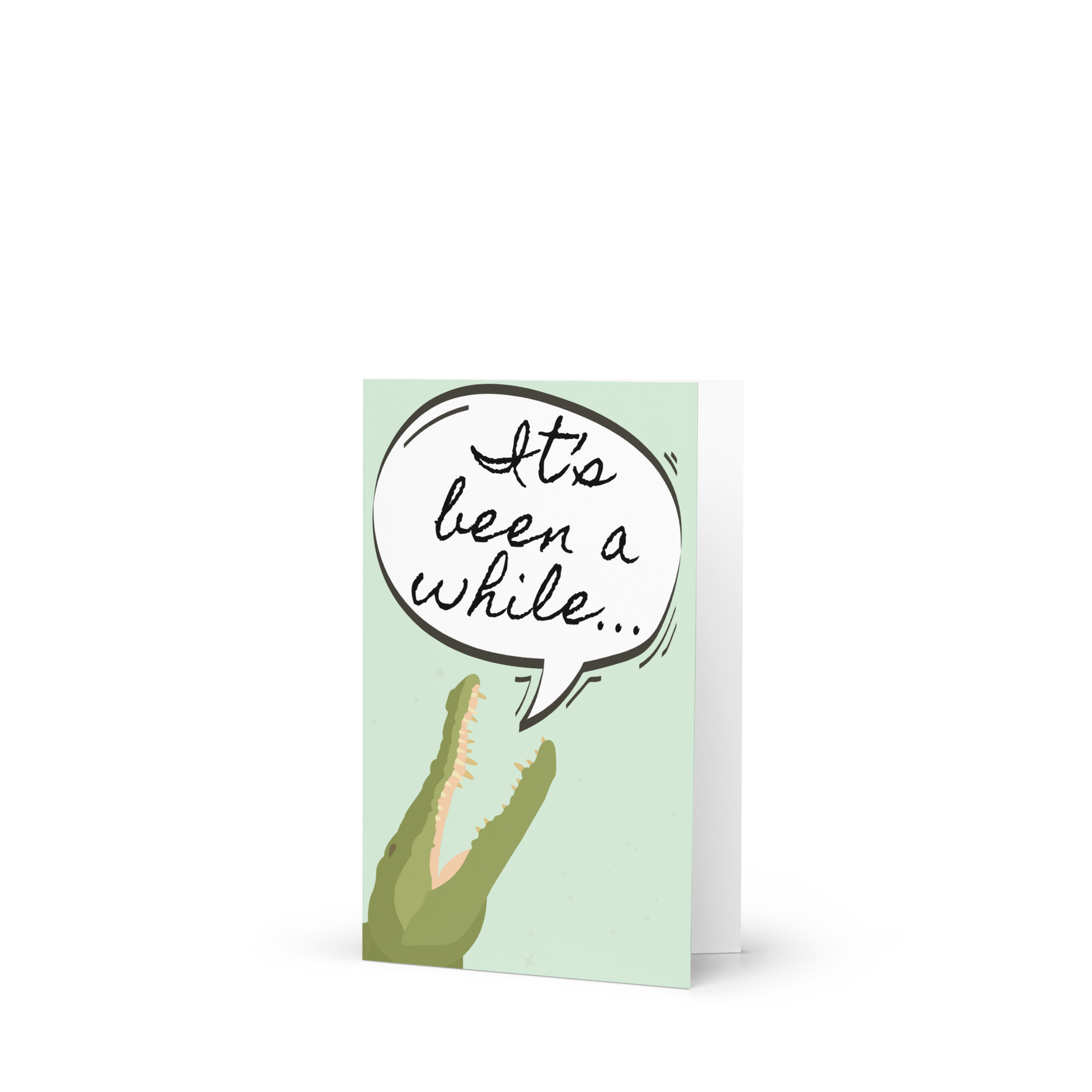 Greeting card with a crocodile and speech bubble saying 'It's been a while' on a light green background