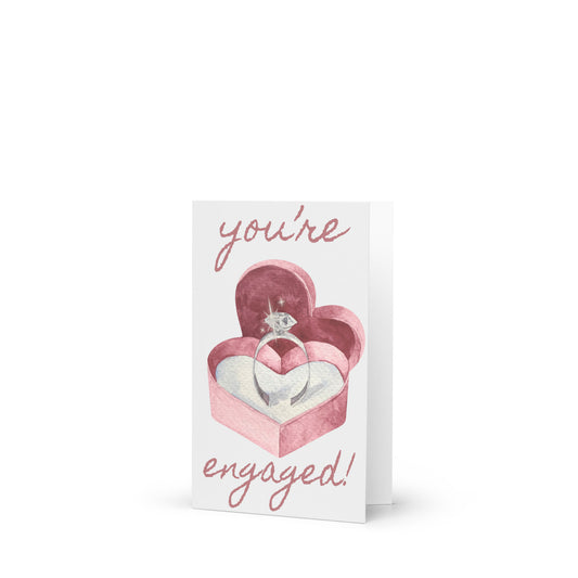 Greeting card with a heart-shaped box containing an engagement ring and 'you're engaged!' text on a white background.