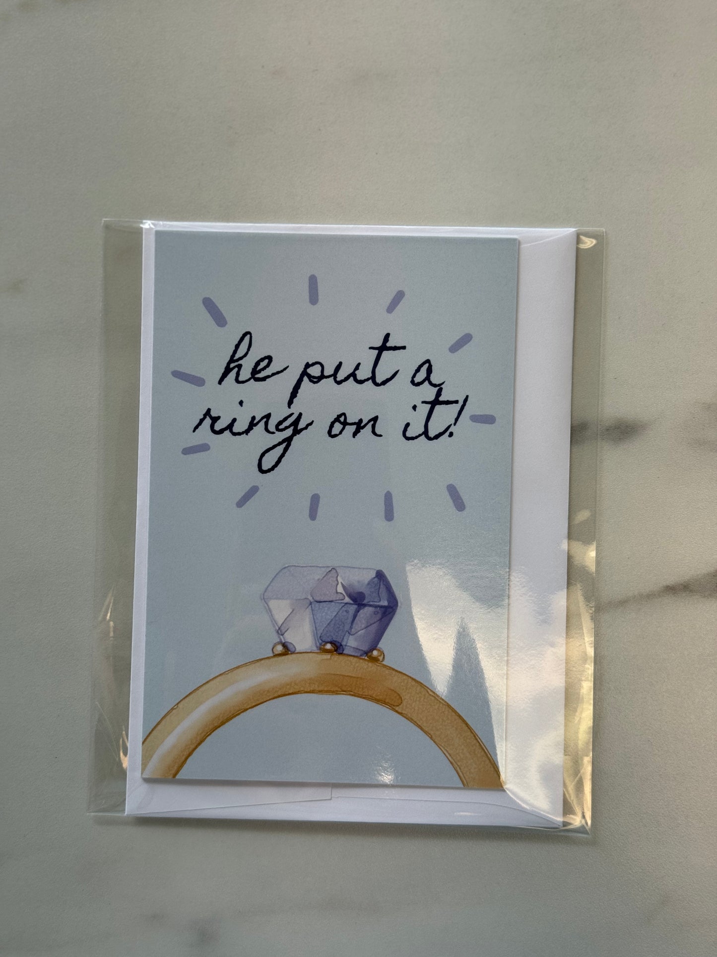 Greeting card with a diamond ring illustration and text 'he put a ring on it!' on a light gray background.